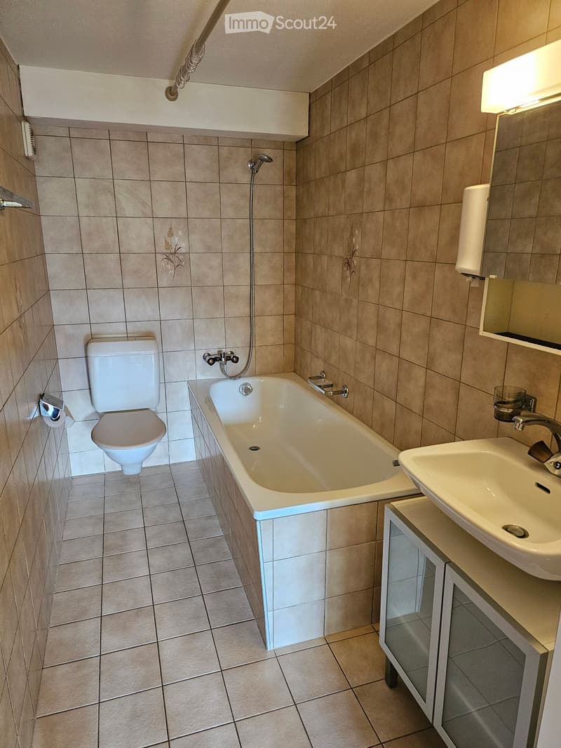 Bathroom with beige tiled walls and floor, featuring a bathtub, toilet, sink, and mirror. Showerhead and faucet are visible.