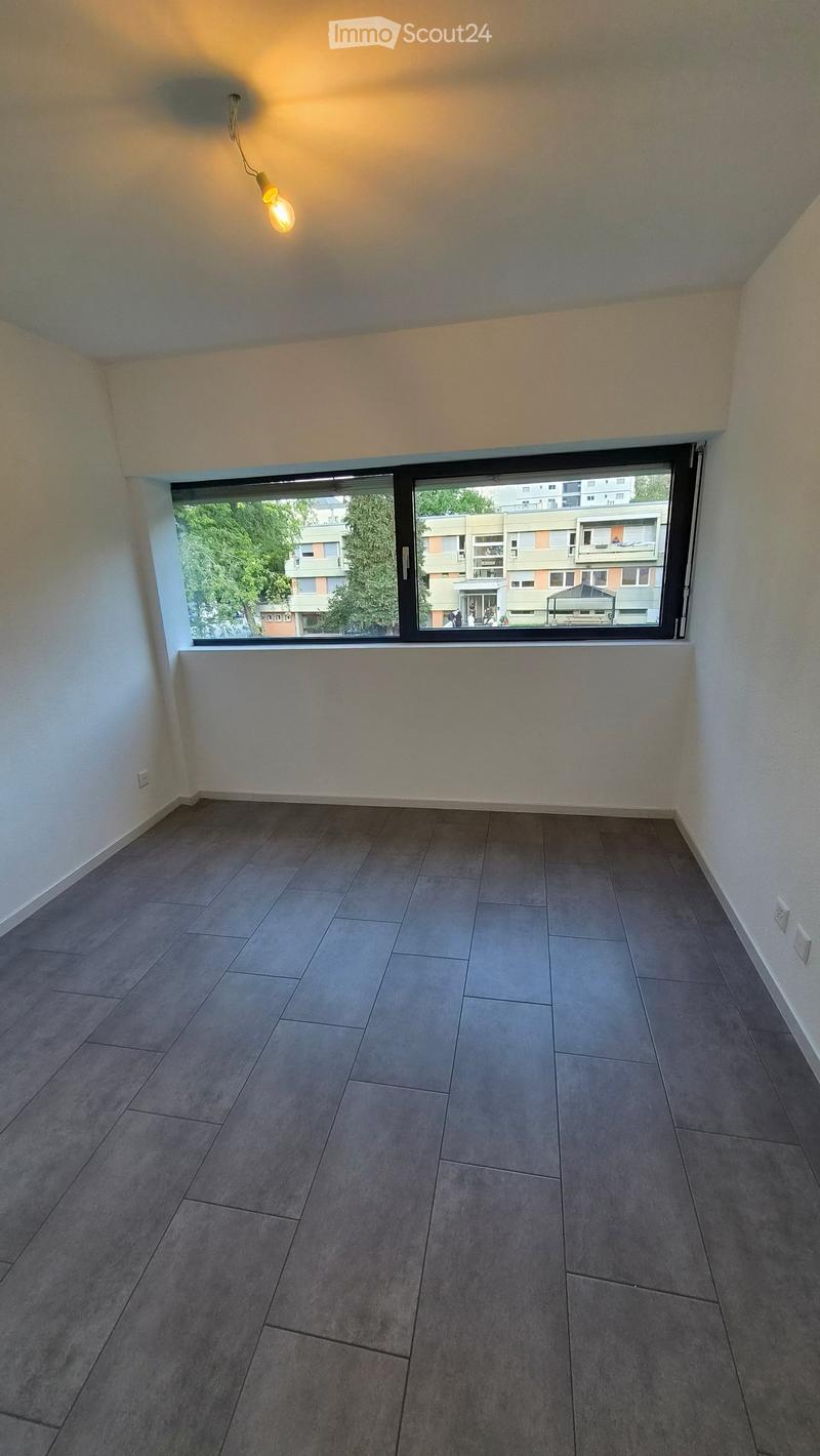 Empty room with grey tiled floor, white walls, large window with view of buildings and trees, and a single hanging light fixture.