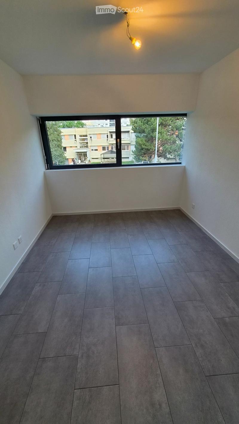 Empty room with grey tiled floor, white walls, large window with view of buildings and trees, and a single hanging light fixture.