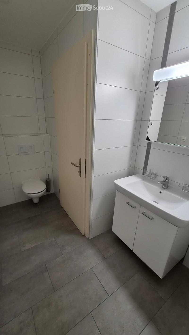 Modern bathroom with white tiles, a toilet, a sink with a mirror, and a small cabinet. The floor is tiled in a dark gray color.