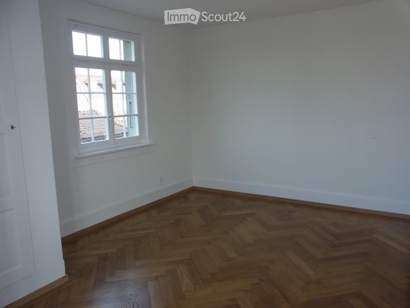Empty room with wooden herringbone flooring, white walls, and a large window with white frame showing a view of rooftops.