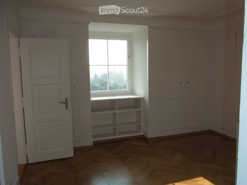 Empty room with wooden flooring, white walls, built-in shelving unit, and a window with a view of trees and a church spire.