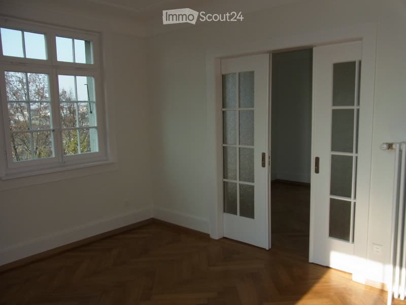 Empty room with wooden flooring, white walls, large windows, and a white door leading to another space.