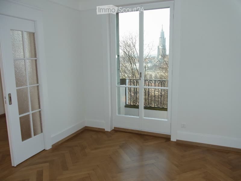 Empty room with wooden herringbone flooring, white walls, and a large window offering a cityscape view.