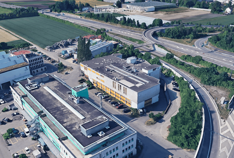 Industrial buildings in a suburban area, surrounded by trees and vegetation, featuring multiple parking areas and access roads.
