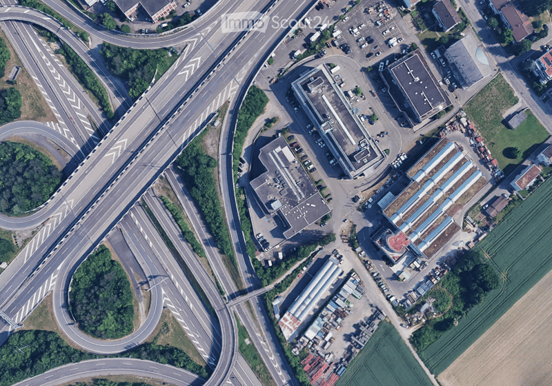 Highway with multiple lanes, buildings on the side, parking lot with several cars, trees, and plants, fields