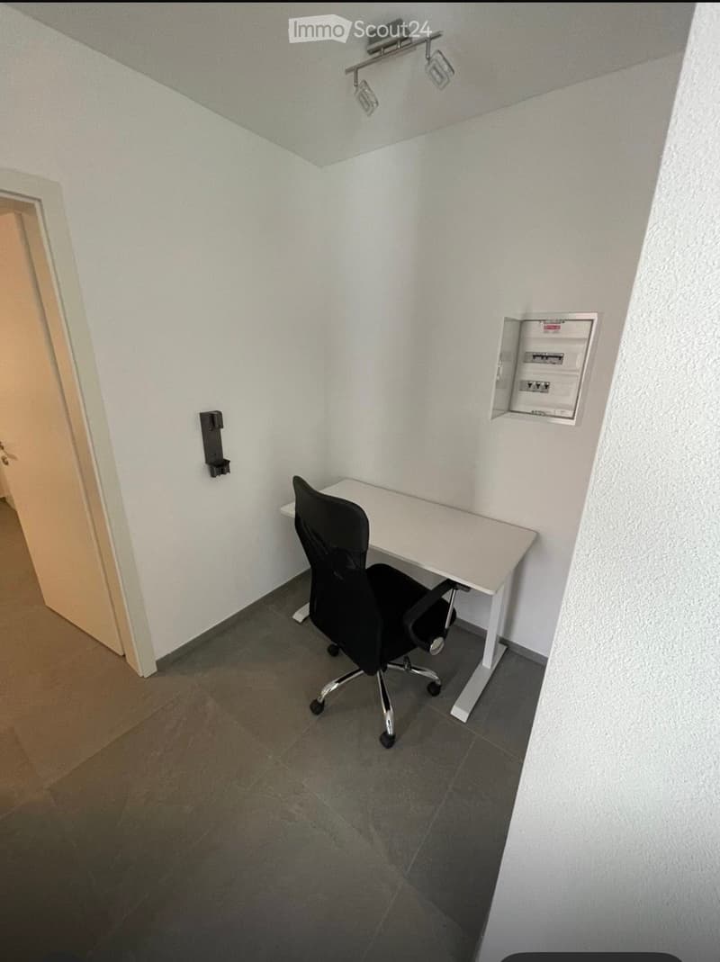 A small, furnished room with a white desk, black office chair, and modern lighting fixture on the ceiling.