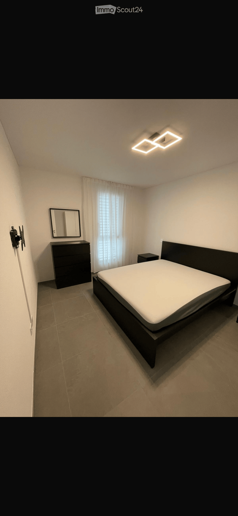 Modern bedroom with a large bed, nightstand, dresser, and mirror. The room features light-colored walls, tiled flooring, and a window with white curtains.