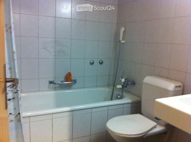 A small bathroom with a bathtub, toilet, sink, and a showerhead attached to the wall. The walls and floor are tiled in light gray.