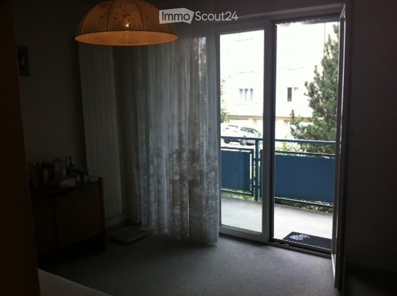 A small, furnished room with a sliding glass door leading to a balcony, a side table with items, a curtain, and a hanging lamp.