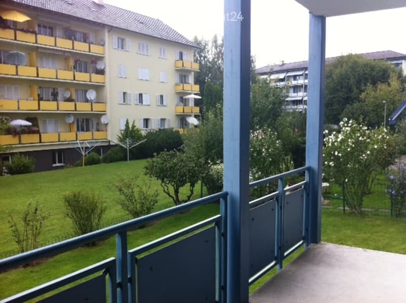 Balcony overlooking a green courtyard with trees and shrubs, featuring a metal railing and a view of neighboring apartment buildings with yellow balconies.