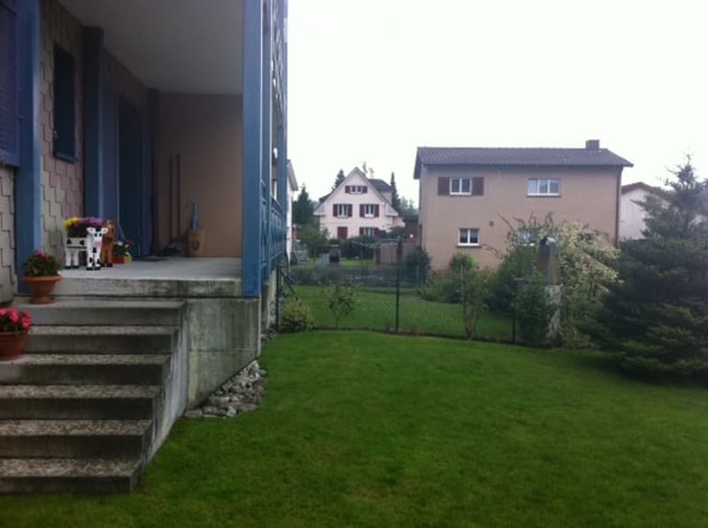 A well-maintained garden with a green lawn, a small porch with potted plants, and a staircase leading up to the house entrance. The background shows neighboring houses and a fence.