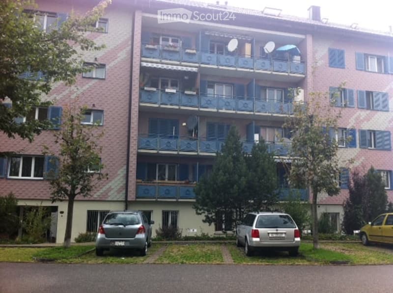 Multi-story apartment building with balconies, satellite dishes, and a parking area with several cars.