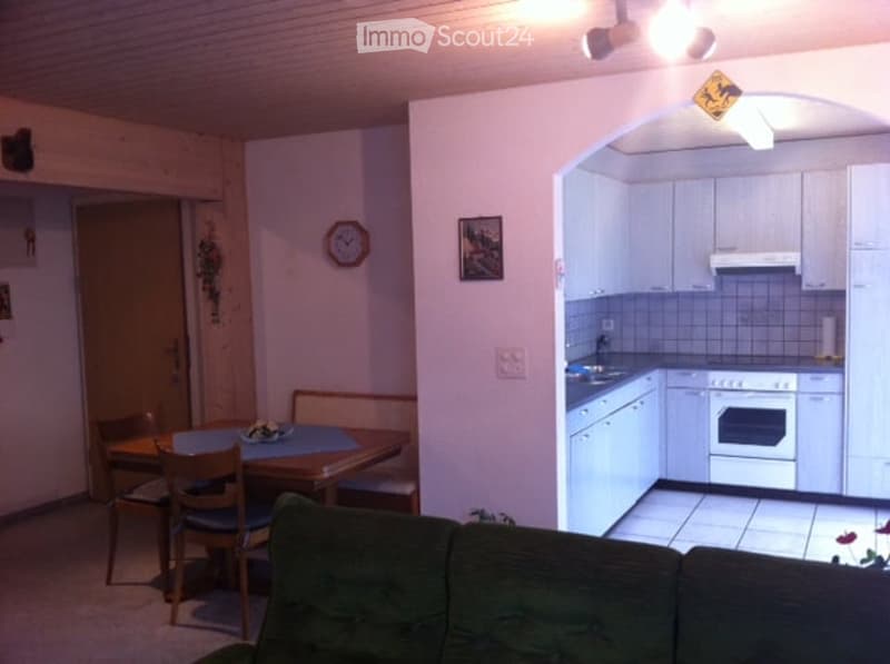 A cozy, open-plan living and dining area connected to a compact kitchen. The living area features a green sofa, while the dining area includes a wooden table with chairs. The kitchen has white cabinets, a stove, and an oven.