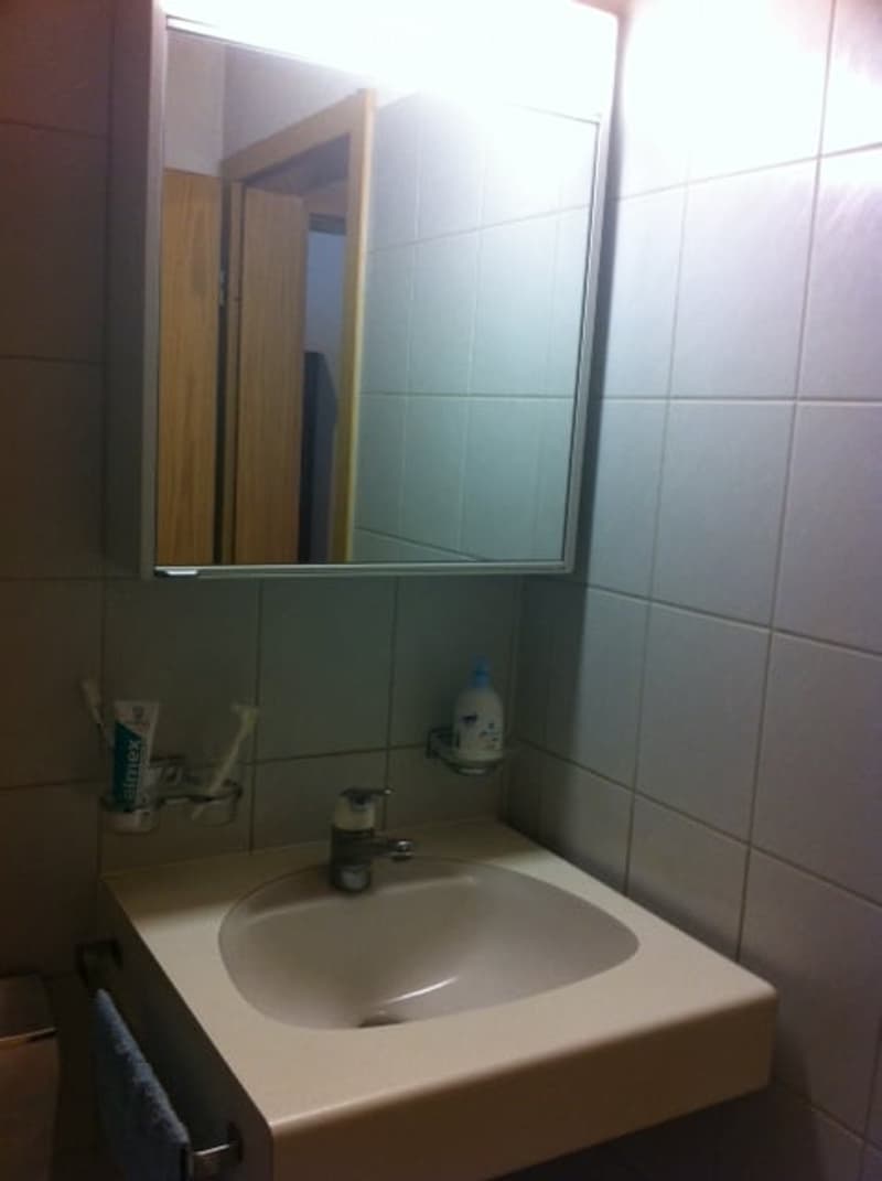 A small bathroom with a white sink, a mirror above it, and a toothbrush and toothpaste on the counter. The walls are tiled in light blue.