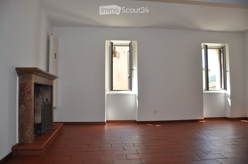Empty room with terracotta tiled floor, white walls, fireplace, and two open windows letting in natural light.