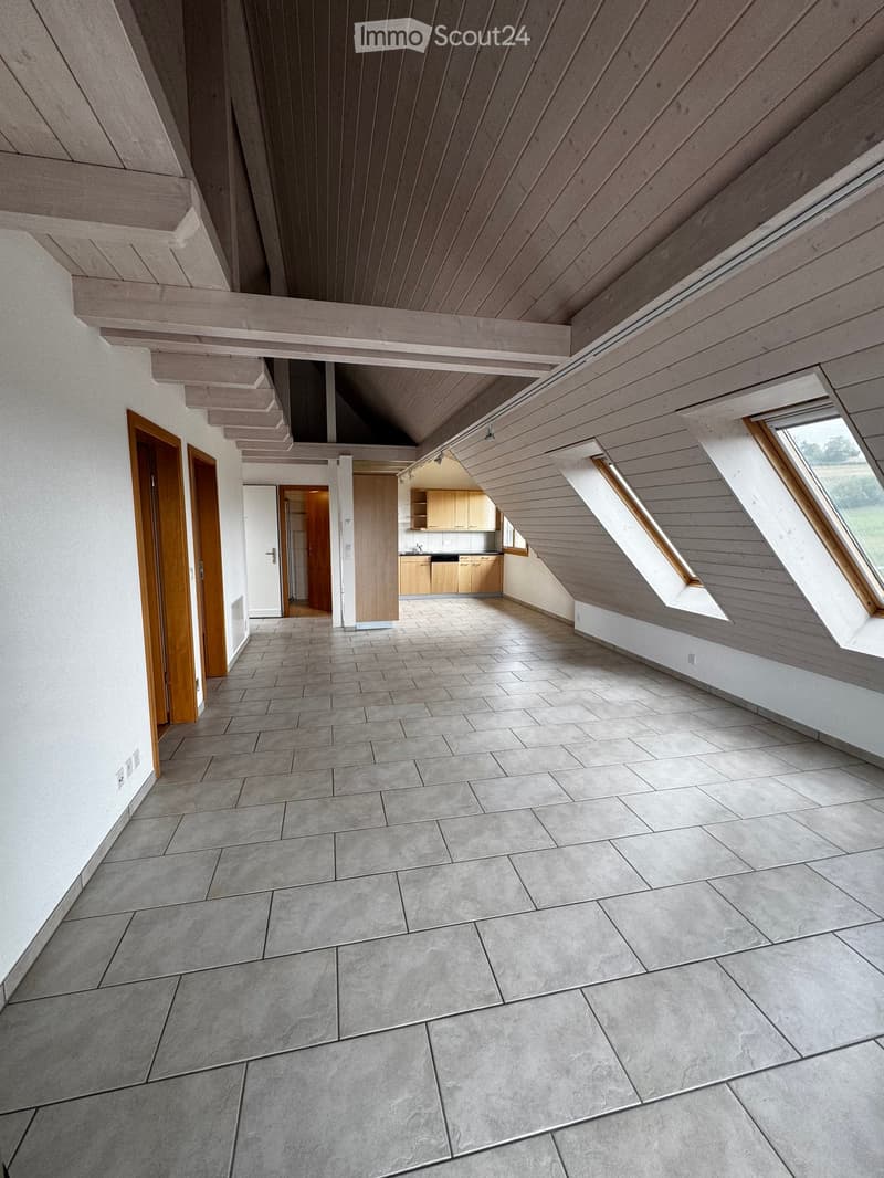Open, modern hallway with grey tiled flooring, wooden ceiling beams, and large skylight windows. Kitchen area visible in the background with light wood cabinets and a sink.