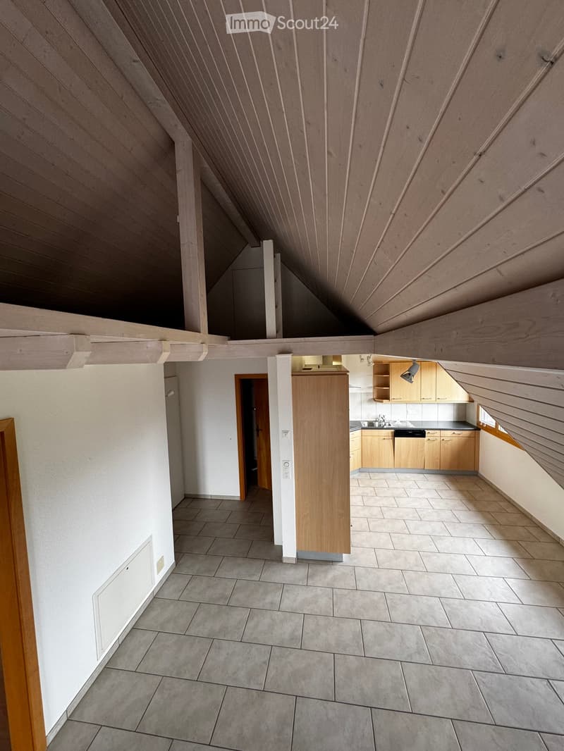 An unfurnished attic space with exposed wooden beams, tiled floor, and a small kitchen area visible in the background.
