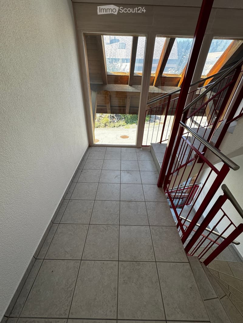 Hallway with tiled floor, red metal staircase railing, and large windows leading to an outdoor area.