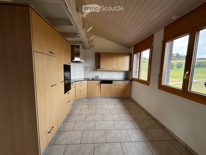 Modern kitchen with light wooden cabinets, tiled floor, and large windows overlooking a green field.