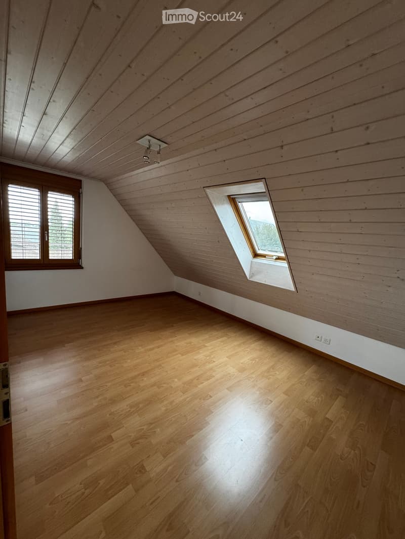 Empty attic room with wooden floors and slanted wooden ceilings, featuring two windows and a door.