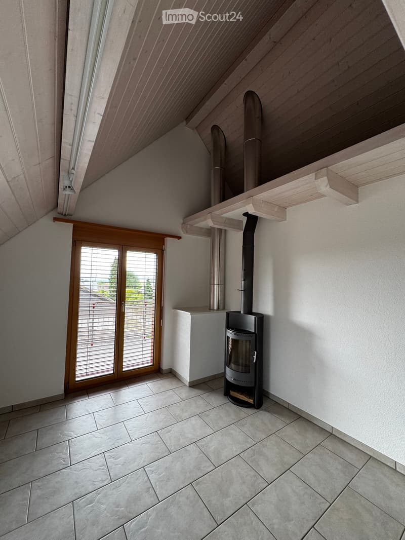 An unfurnished attic room with a modern wood-burning stove, large skylight, and wooden shelving unit. The room features white walls, a tiled floor, and a sliding glass door leading to a balcony.