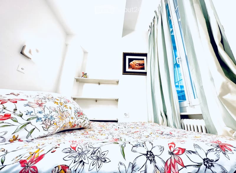 A cozy bedroom with a bed covered in a floral-patterned duvet and pillow, a window with light-colored curtains, a framed picture on the wall, and a small shelf with decorative items.