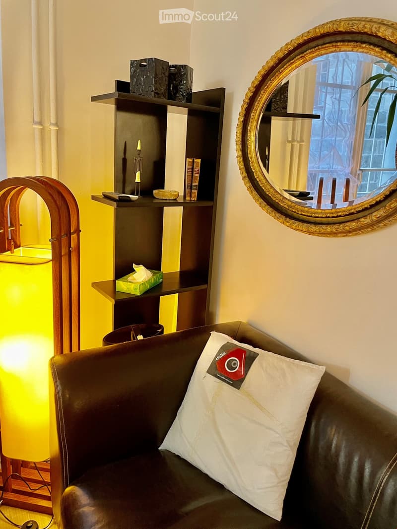 A cozy living room with a dark leather sofa, a large ornate mirror, a wooden shelf with books and decorative items, and a bright yellow lamp.