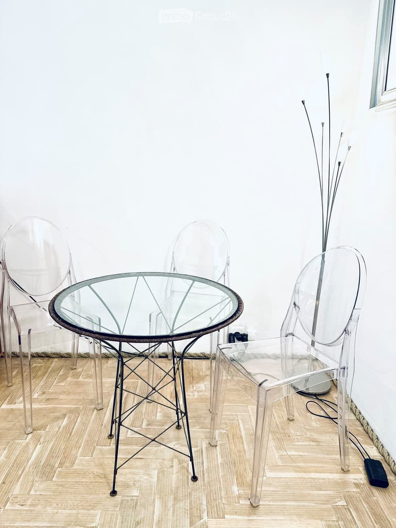 A modern, minimalist room with a glass table and four transparent chairs, wooden herringbone flooring, and white walls.