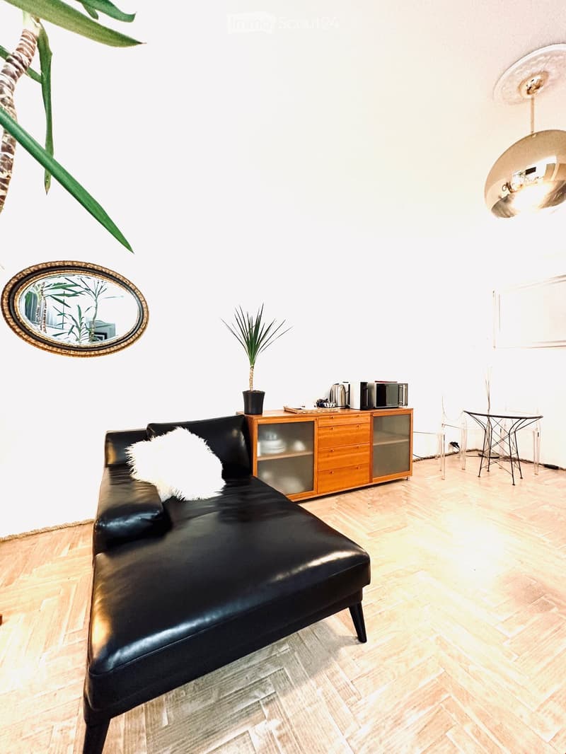 A modern living room with a black leather sofa, a wooden cabinet, a small table, a chair, a decorative mirror, a potted plant, and a hanging lamp.