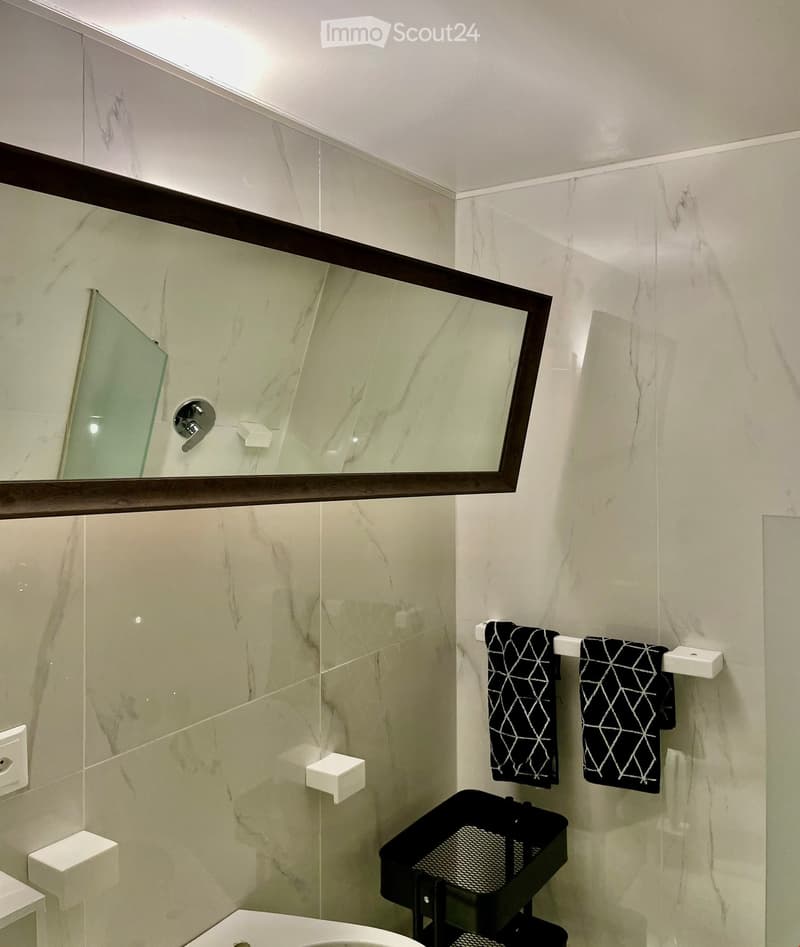 Modern bathroom with marble tiles, a large mirror with wooden frame, towel rack with two black towels, and a toilet.