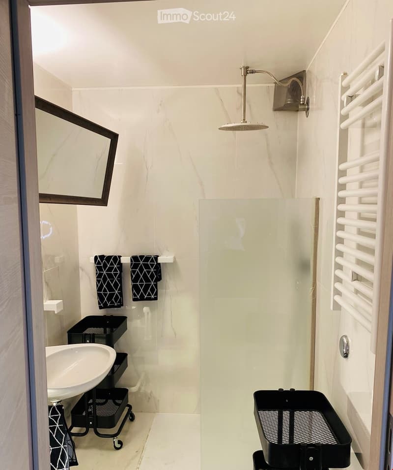 Modern bathroom with white marble tiles, a glass-enclosed shower, a sink with a mirror, and a radiator. Two black towels are hanging on the wall.
