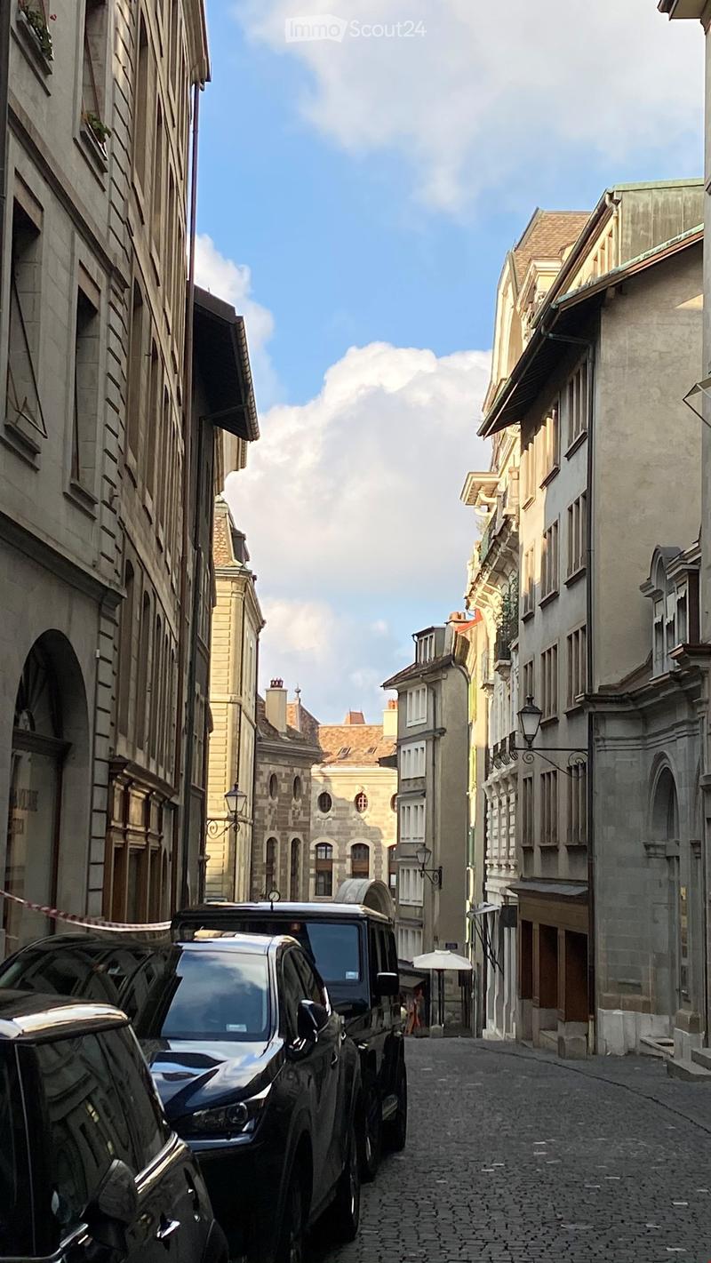A narrow cobblestone street lined with multi-story buildings, featuring classic European architecture with arched windows and balconies. A parked vehicle is visible on the left side.