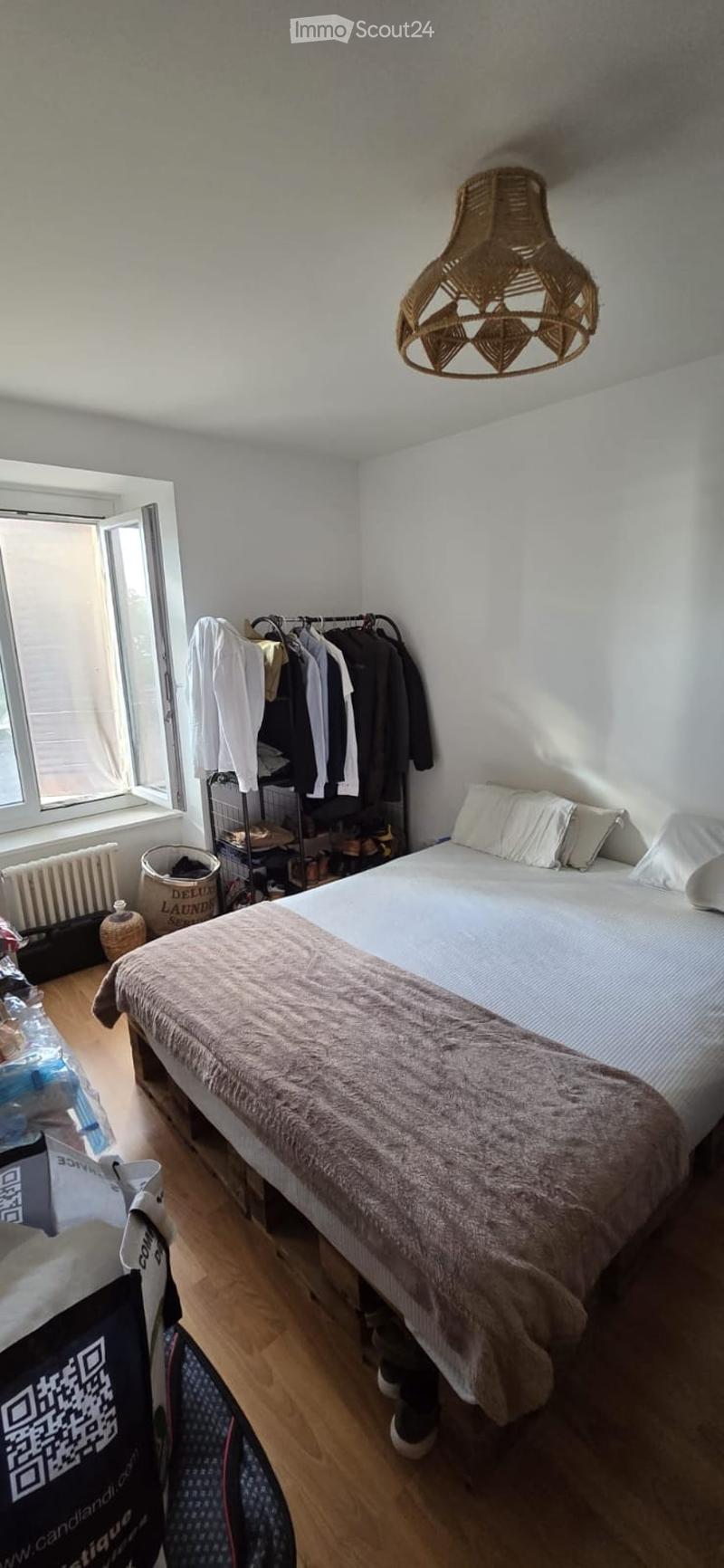 A cozy bedroom with a double bed, pillows, and a blanket. A wardrobe with hanging clothes is visible, along with a radiator, a wicker basket, and a stylish wicker pendant light.