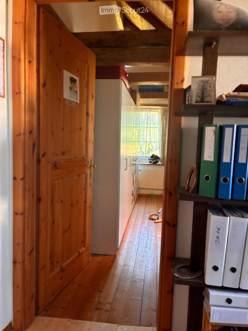 Wooden hallway with open door leading to another room, visible wooden floor, shelves with binders, and a broom in the adjacent room.