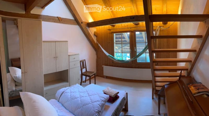 Cozy attic bedroom with wooden beams, a bed, a chair, a wardrobe, a hammock, and a staircase leading to another level.