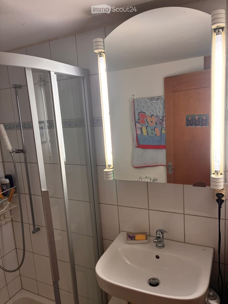 Bathroom with a sink, faucet, shower, towel, and soap dish. The walls are tiled, and there is a wooden cabinet with a mirror above the sink.