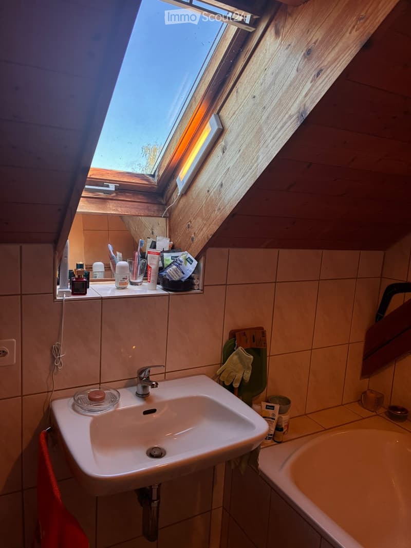 Bathroom with a sink, mirror, and bathtub, featuring wooden slanted ceilings and a skylight window. Personal care items are placed on the sink counter.