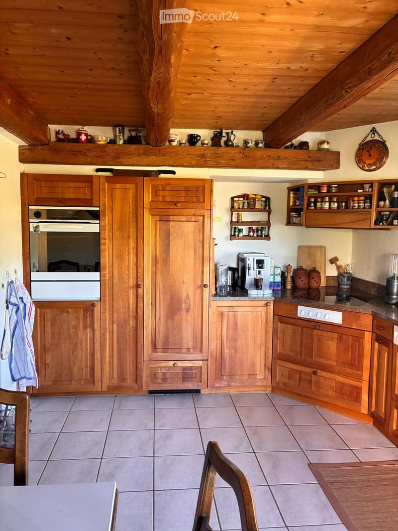 Cozy kitchen with wooden cabinets, tiled floor, and various appliances including an oven, coffee maker, and blender. Shelves hold spices and decorative items, and a dining area with a table and chairs is visible.