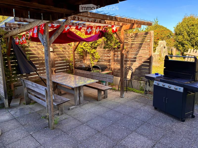 Wooden pergola with a table, benches, chairs, and a grill, surrounded by a wooden fence and greenery.