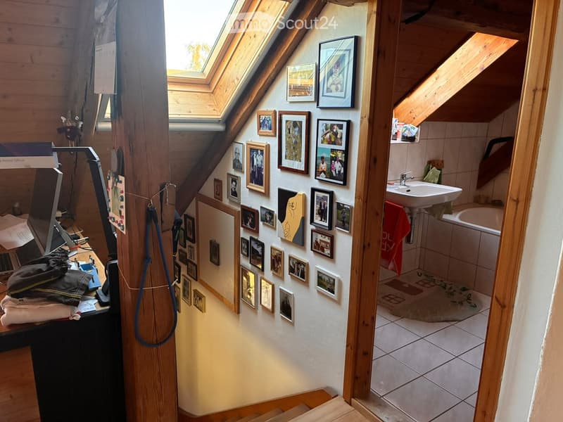 Loft-style room with wooden beams, skylight windows, and a small bathroom area visible through an open doorway. The room features a desk with a chair, framed pictures on the wall, and a towel hanging on the side.