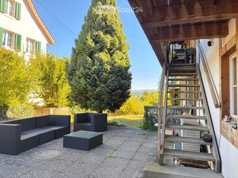 Outdoor patio with a black wicker sofa, matching armchair, and a glass-top coffee table. Wooden stairs lead up to a wooden deck area with additional seating and storage. The background features a house with green shutters and a lush, green landscape.