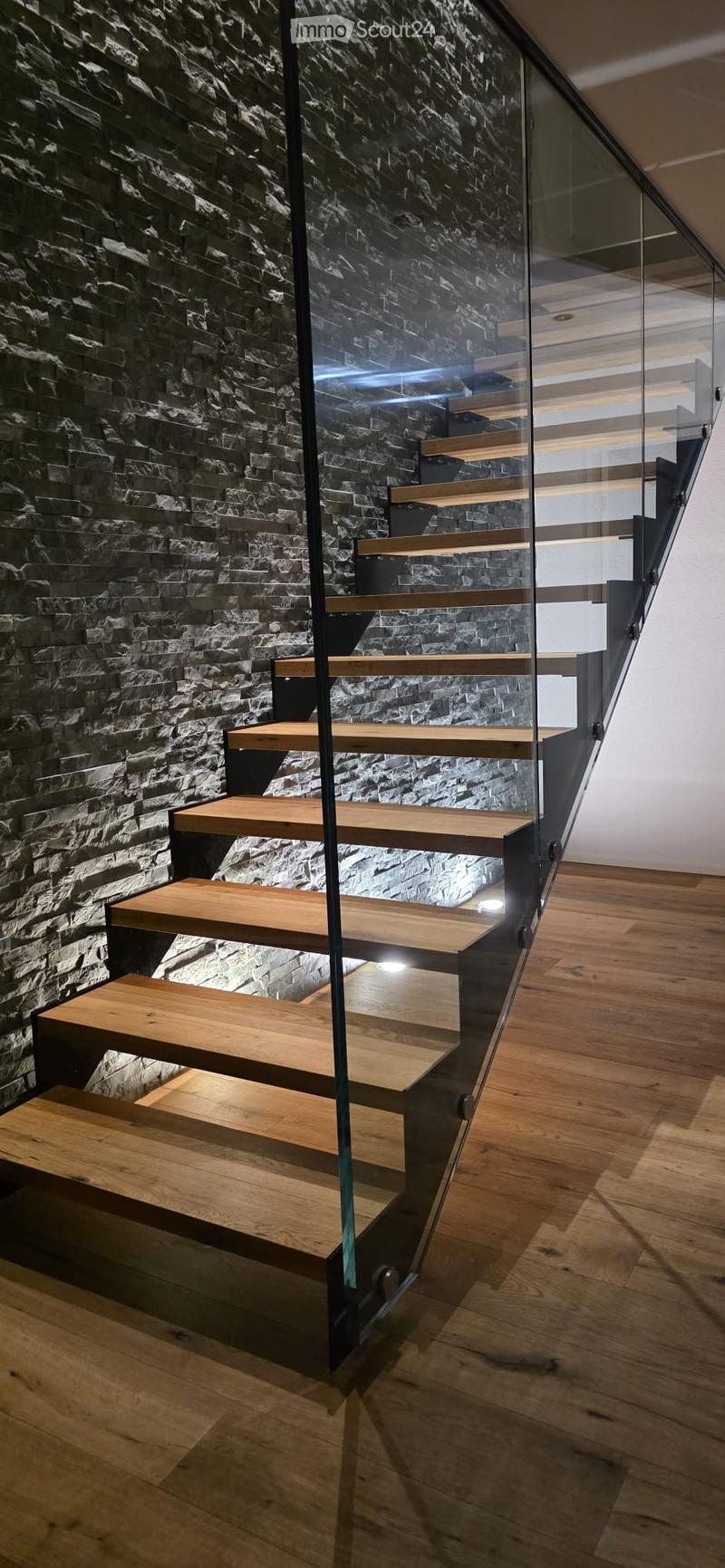 Modern staircase with wooden steps and glass railing, set against a textured stone wall, leading to an upper level with wooden flooring.