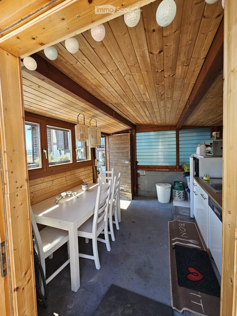 A cozy attic space with wooden walls and ceiling, featuring a dining area with a white table and chairs, a small kitchen area with a refrigerator, sink, and countertop, and several hanging pendant lights.