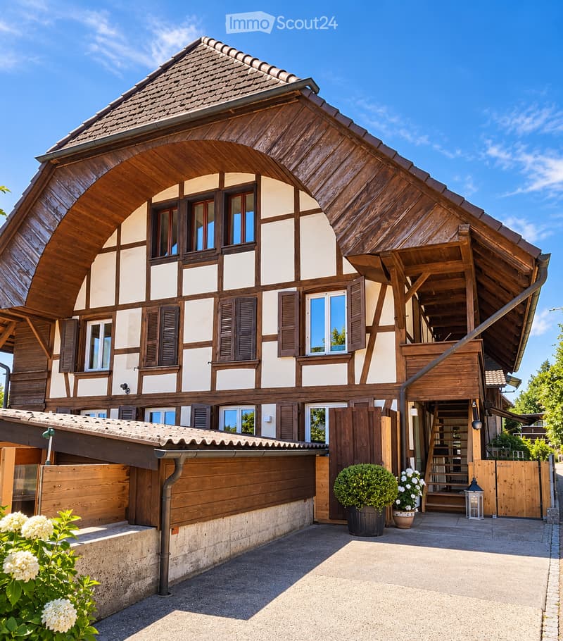 Traditional wooden house with a steeply pitched roof, white and brown facade, multiple windows with shutters, and a covered entrance area with steps leading up to the door.