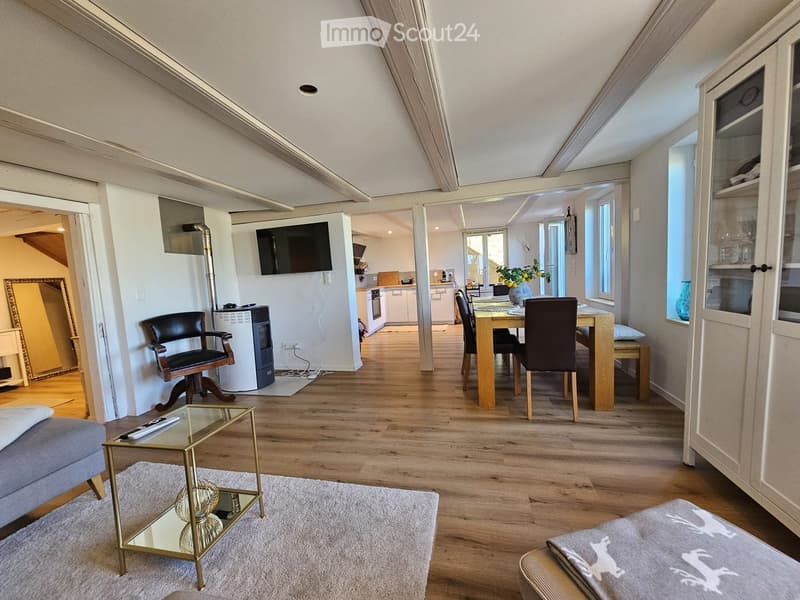 A modern, open-concept living and dining area with wooden flooring, a grey sofa, a glass coffee table, a dining table with chairs, a television mounted on the wall, and a small kitchen area visible in the background.