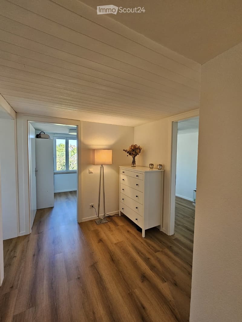 A narrow hallway with wooden flooring, white walls, and a slanted ceiling. A small white drawer unit with a vase of flowers sits near a tall table lamp. The hallway leads to an open area with large windows and a view of greenery.