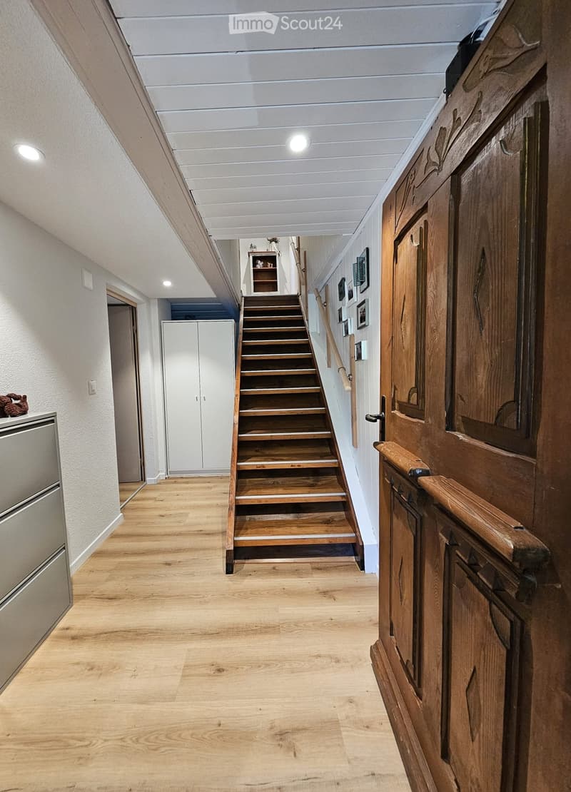Spacious hallway with wooden flooring, white walls, and a staircase leading upwards. Features built-in cabinets, framed artwork, and recessed lighting.