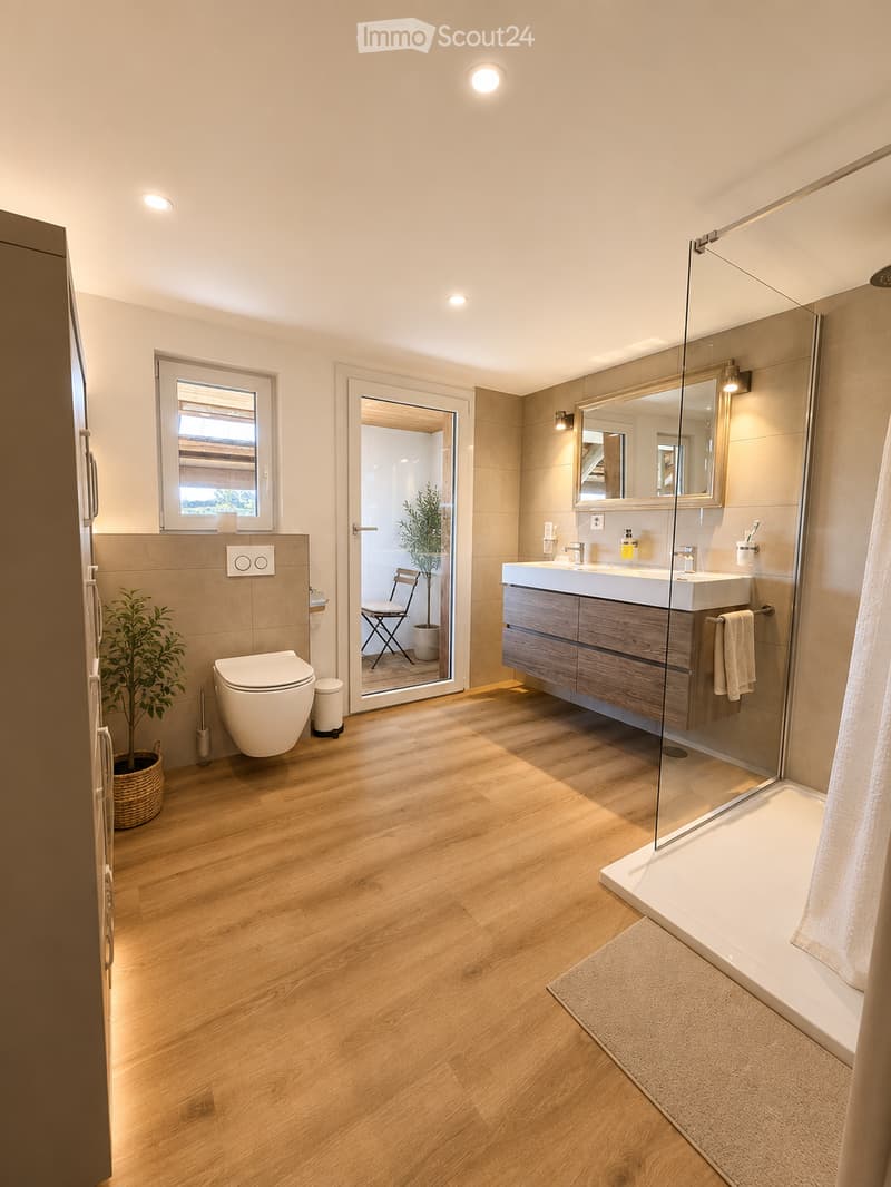 Modern bathroom with wooden flooring, a toilet, a double sink with a wooden cabinet, a large mirror, a glass-enclosed shower, and a small window. A potted plant is placed near the toilet, and a chair is visible through the open door leading to another roo