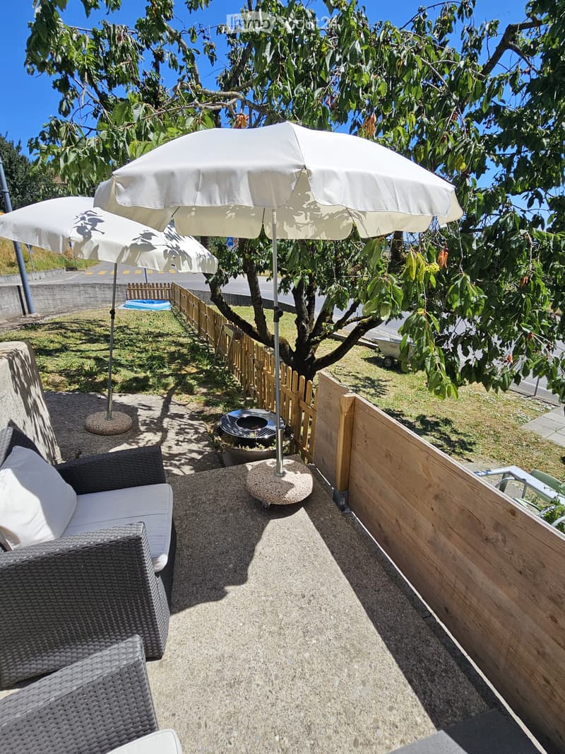 Balcony with a wicker sofa, large umbrella, and a small table under a tree.